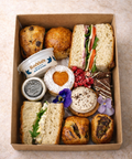 Assorted food items including sandwiches, pastries, and tarts in a cardboard box on a light background.